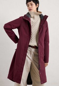 Bordeauxkleurige parka met een gestructureerde grijze kraag, rits- en drukknoopsluiting, zijzakken, gedragen over een crèmekleurige kabelgebreide trui en lichte corduroybroek.