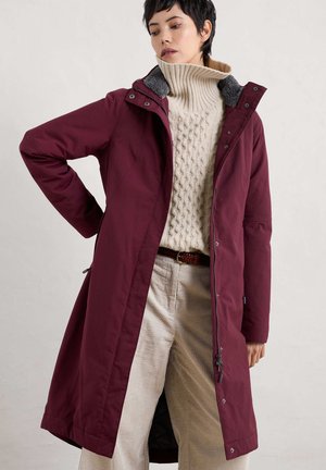 Parka bordeaux avec un col texturé gris, fermeture éclair et pression, poches latérales, portée sur un pull en tricot torsadé crème et un pantalon en velours côtelé clair.