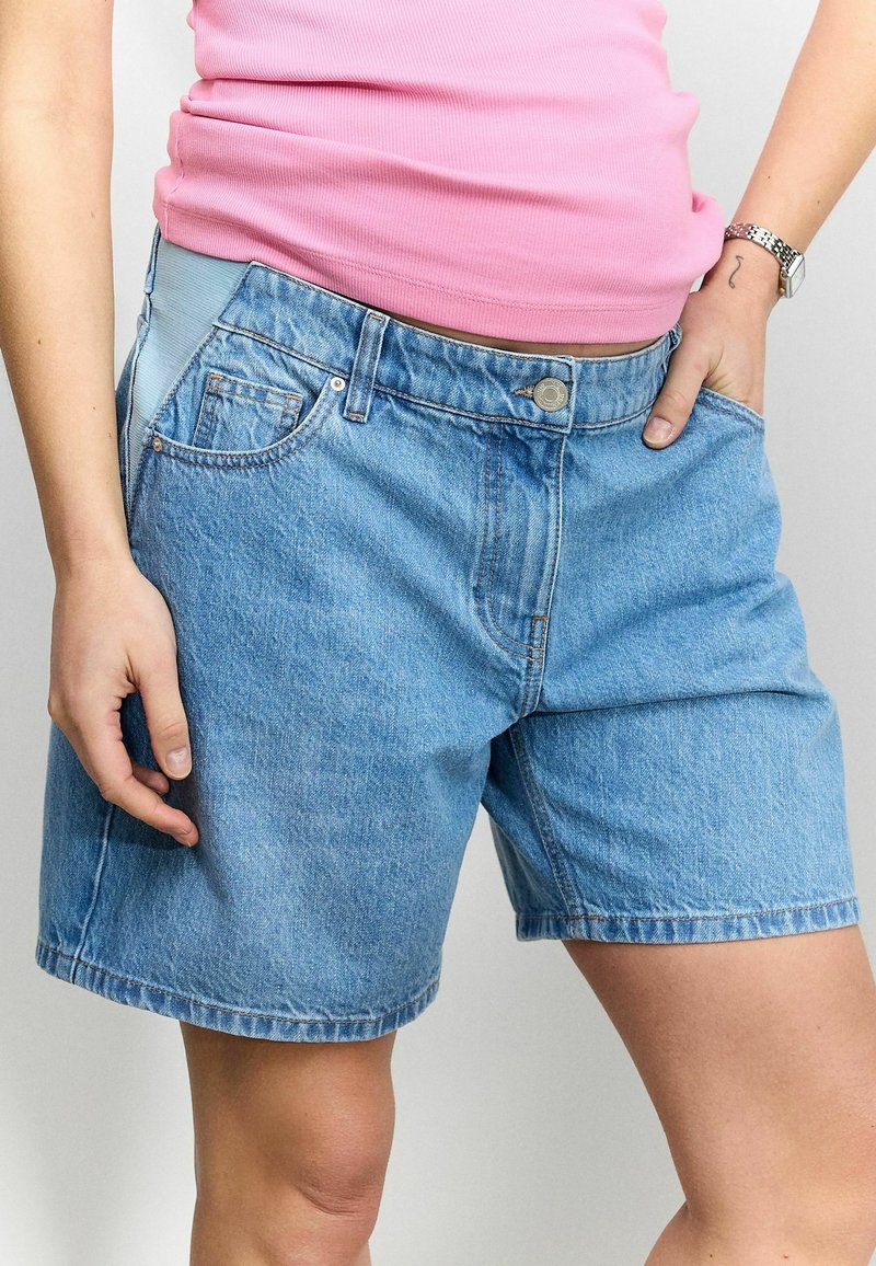 Femme portant un haut rose et un short en jean bleu de maternité avec un panneau de taille élastique, posant avec les mains près des poches.