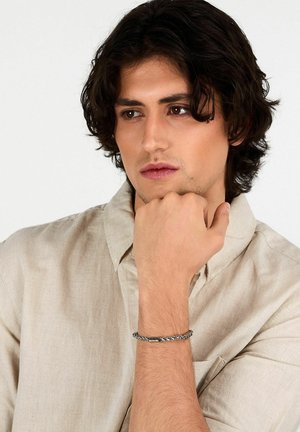 Braccialetto in catena d'argento con maglie rettangolari, indossato su un polso leggermente abbronzato, abbinato a una camicia beige con bottoni.