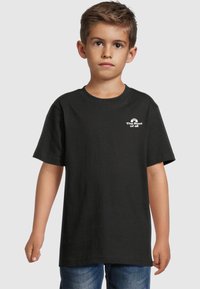 Juodas medvilninis trumpomis rankovėmis t-shirt, su apvaliu kaklu ir mažu baltu grafiniu tekstu logotipu kairėje krūtinės pusėje. Kasdienė išvaizda.
