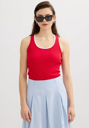 Mujer con gafas de sol negras, top sin mangas rojo con ribete blanco y falda plisada azul claro, posando sobre un fondo liso.