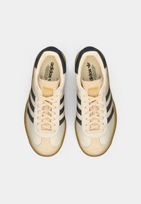 Beige and navy athletic shoes with a suede and textile upper. Features three navy stripes, cream laces, and a gum rubber sole.