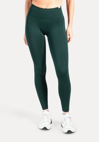 Leggings verde scuro a vita alta realizzati in tessuto elasticizzato. Presentano una fascia in vita a coste e pannelli laterali testurizzati. Indossati con sneaker bianche.
