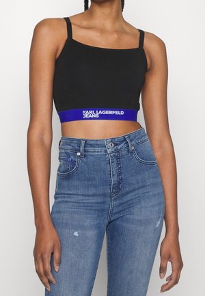 Femme portant un débardeur noir court avec l'inscription "KARL LAGERFELD JEANS" sur une bande bleue et un jean en denim bleu taille haute.