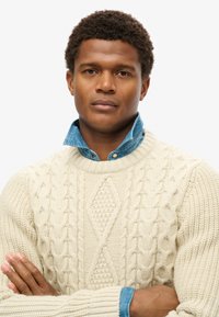 Cremfarbener, kabelgestrickter Pullover mit strukturiertem Muster, über einem himmelblauen Jeanshemd mit spitzem Kragen getragen.