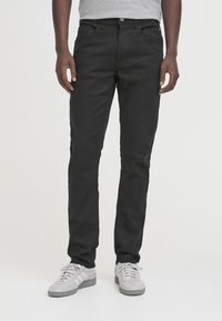 Pantaloni neri a taglio slim in misto cotone, con classico design a cinque tasche e chiusura con bottone. Indossati con scarpe da ginnastica grigie.