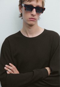Junge Person mit hellbraunen Haaren, die dunkle rechteckige Sonnenbrille und einen dunkel gestreiften Pullover trägt, mit verschränkten Armen vor einer weißen Wand.