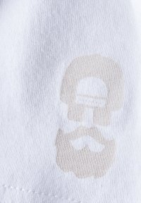 Weißer Stoff mit einem hellbeige Druck eines Schädels mit einem Schnurrbart. Die Textur ist glatt und hat ein feines Strickmuster.