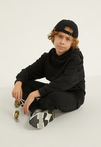 Melnā kapuche un sporta bikses, kas izgatavotas no mīksta auduma. Zēns sēž ar kājām pāri, uzvelkot melnu cepuri un turot atstarojošas saulesbrilles. Pelēki sporta apavi.