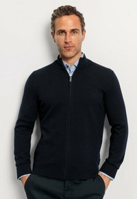 Navy zip-up sweater with a stand collar, knit texture, and ribbed cuffs. Worn over a light blue collared shirt for layering.