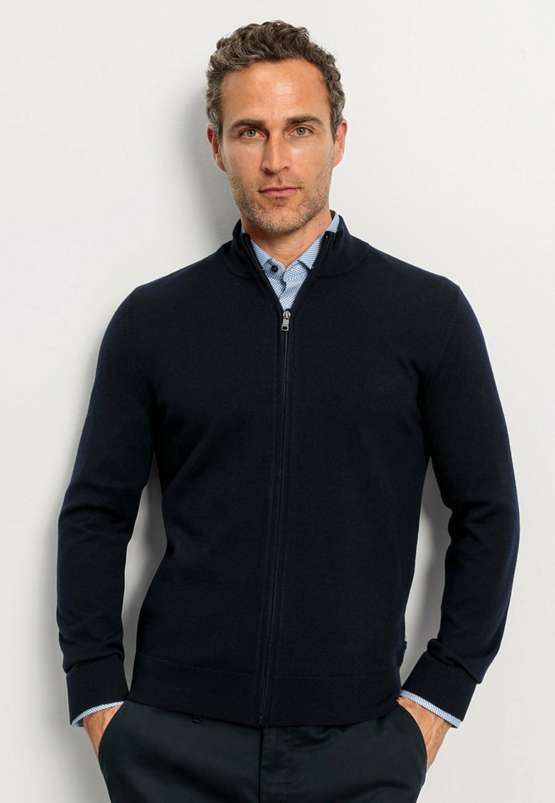 Navy zip-up sweater with a stand collar, knit texture, and ribbed cuffs. Worn over a light blue collared shirt for layering.