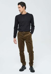 Sweater noir en tricot, pantalon en velours côtelé marron avec une taille à cordon, et bottes noires à lacets. Le mannequin se tenait devant un fond uni.