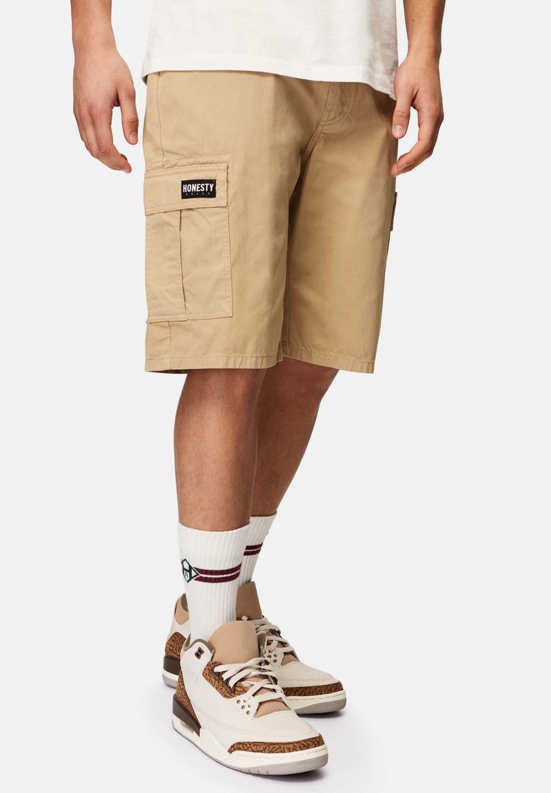 Beige cargo shorts med to sidelommer, syet detaljer og en sort "HONESTY" label. Matchet med hvide høj-top sneakers og stribede sokker.