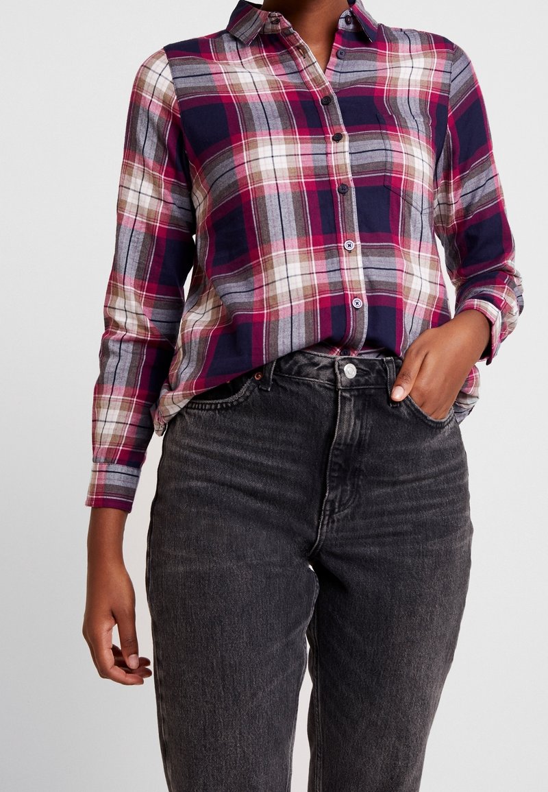Plaid button-up shirt with dark navy, pink, gray, and cream colors. Features a collar and long sleeves, paired with high-waisted black jeans.