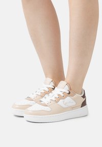 Beige och vit sneaker gjord av läder och syntetiska material, med en vadderad krage, perforerad tå och bruna accenter. Gummisula.