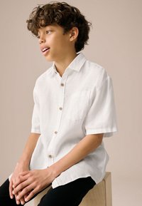 Weißes Leinenhemd mit kurzen Ärmeln, durchgehendem Knopfverschluss, Brusttasche und Holzknöpfen. Das Modell hat Locken und sitzt auf einem Holzblock.