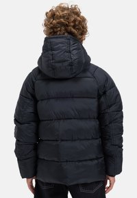 Barn med lockigt hår som bär en svart puffjacka med huva, sedd bakifrån mot en enfärgad vit bakgrund.