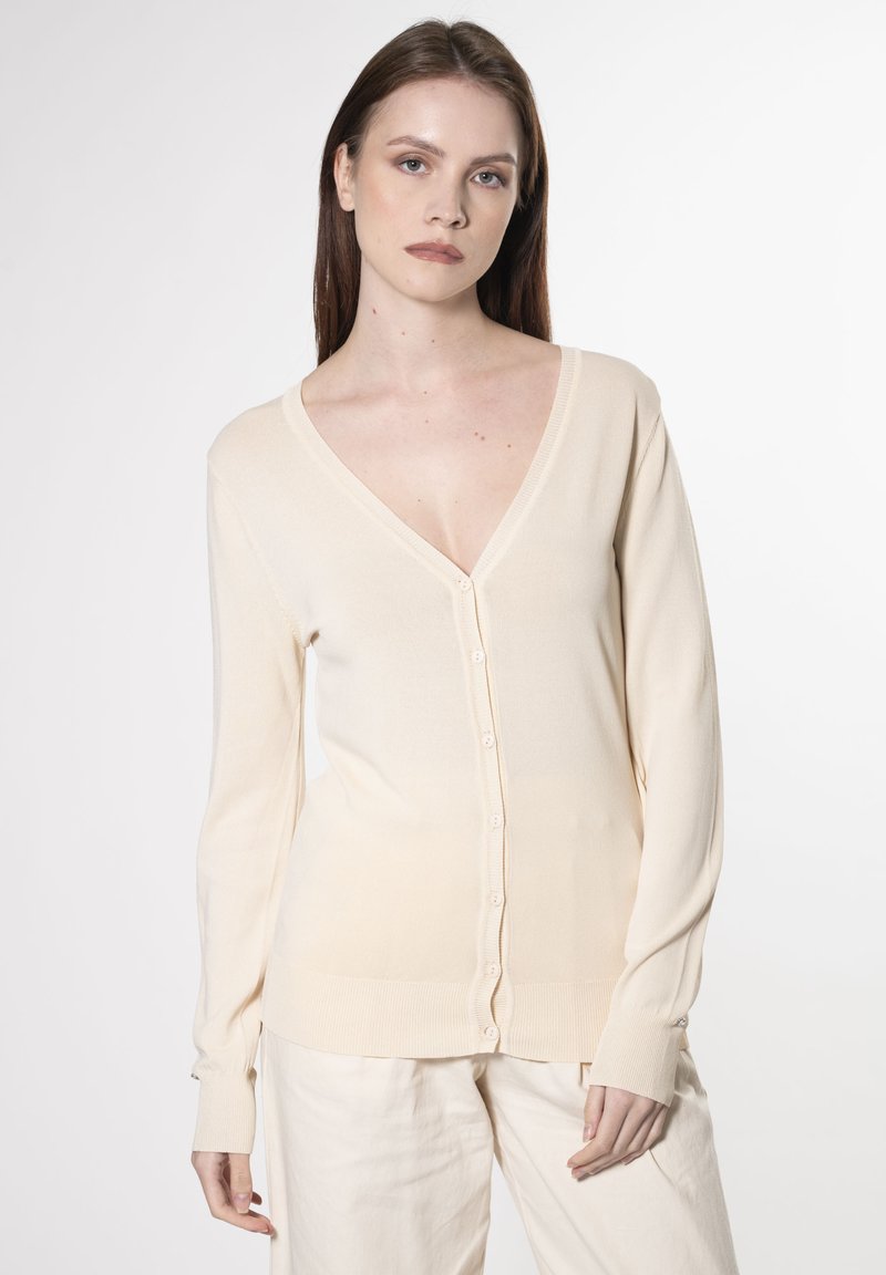 Lys beige cardigan lavet af blødt stof med v-hals, lange ærmer og knaplukning. Har ribbede manchetter og lynlås.