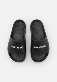 Chanclas de goma negras con diseño de punta abierta. Presentan un patrón de cuadros en relieve en la suela y el logotipo "LACOSTE" en blanco en la parte superior.