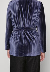 Marineblauwe geplooide fluwelen blazer met lange mouwen, een ceintuur in de taille en een soepele textuur; heeft verticale lijnen en een relaxte pasvorm.