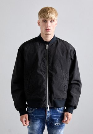 Blonder Mann in einer schwarzen Bomberjacke, die zugezippt ist, und einer stark verwaschenen blauen Jeans, steht vor einem einfarbigen hellgrauen Hintergrund.