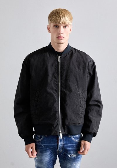 Un homme blond portant une veste bomber noire zippée et un jean bleu déchiré, se tenant devant un fond gris clair uni.