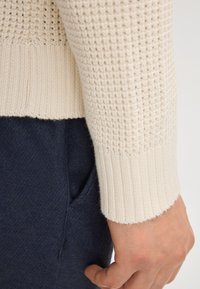 Dettaglio di un maglione lavorato a maglia color crema, con trama testurizzata, polsini a coste e un indumento blu con tasche sotto.