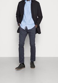 Schwarzer langer Mantel, hellblaues Hemd mit Knöpfen, marineblaue Hose und graue Schuhe mit heller Sohle. Die Hose hat eine glatte Textur; die Schuhe sind aus Wildleder.