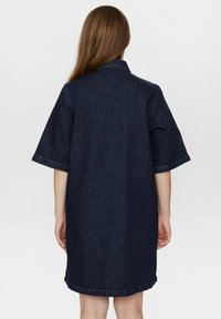 De donkere denim jurk heeft een kraag en wijde mouwen. De zoom is iets gebogen en zichtbare stiksels versterken het ontwerp.