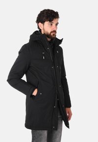 Parka nera imbottita con cappuccio, caratterizzata da chiusura con zip e bottoni a pressione, tasche laterali e corde di regolazione.