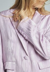 Blazer di un leggero lavanda con un motivo testurizzato e ondulato. Caratterizzato da un colletto a scollatura a V, chiusura con un bottone singolo e bottoni decorativi sulle maniche.