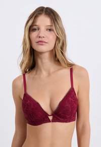 Soutien-gorge en dentelle bordeaux avec un design en V, des bretelles ajustables et un détail en trou de serrure au centre. Des bords festonnés délicats ornent les bonnets.