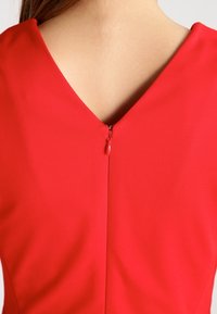Robe rouge avec un décolleté en V et une fermeture éclair dans le dos. Le tissu semble lisse avec une texture unie, mettant en valeur une silhouette épurée et ajustée.