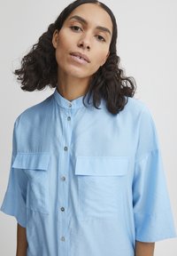 Camicia azzurra a maniche corte con colletto rialzato, due taschini sul petto e chiusura frontale a bottoni. Tessuto morbido e liscio con una leggera lucentezza.