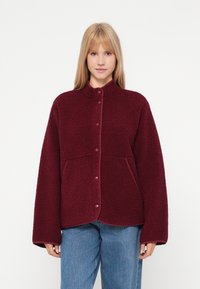 Noor naine kannab burgundi värvi pressnõu ja pikkade varrukatega fliisjakki koos siniste teksadega, taust on ühtlane valge.