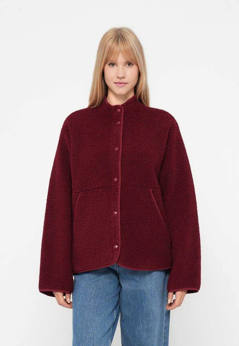 Noor naine kannab burgundi värvi pressnõu ja pikkade varrukatega fliisjakki koos siniste teksadega, taust on ühtlane valge.