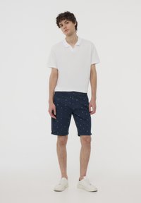 Polo shirt bianco in cotone abbinato a pantaloni corti blu navy con un piccolo motivo a triangolo, risvolti sulle maniche e sneakers chiare.