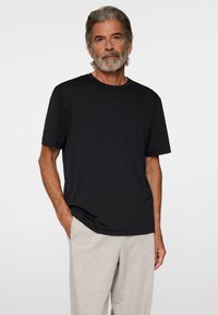 Man med grått hår och skägg som bär en svart t-shirt med kort ärm och ljusgrå byxor, står med en hand i fickan.