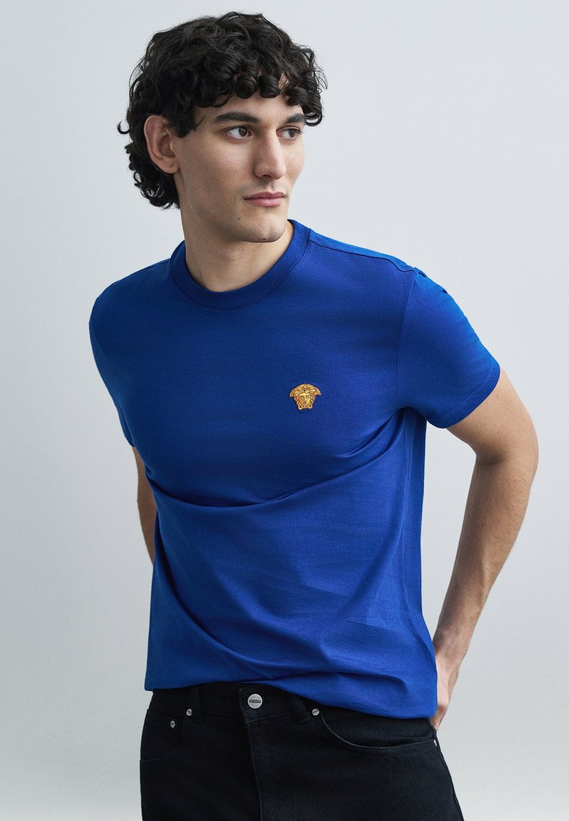 Kék rövid ujjú póló sima anyagból, arany oroszlánfej emblémával a mellkason. Fekete farmerral viselve.