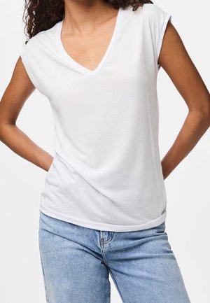 Femme portant un haut blanc sans manches à col en V légèrement rentré dans un jean en denim bleu clair, posant avec les mains derrière le dos.