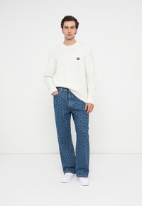 Weißer, strukturierter Strickpullover mit einem Logo-Patch, kombiniert mit blauen, gemusterten Jeans mit ausgestellten Beinen und weißen Sneakers.