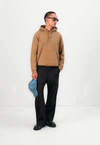 Brown hoodie with a front pocket, black pants, and black shoes. Holding a light blue cap. Sleek sunglasses complete the outfit.