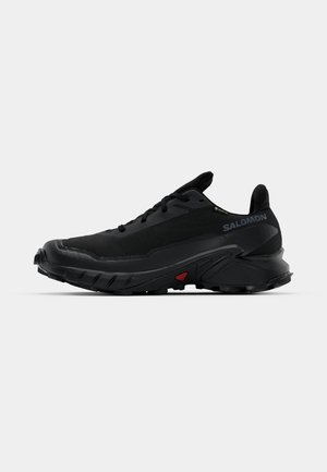 Black athletic shoes with a textured upper, rounded toe, and prominent rubber outsole. Features a logo on the side and red detailing on the sole.