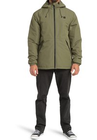 Giacca con cappuccio verde oliva, con zip frontale, polsini elasticizzati e due tasche laterali. Realizzata in materiale liscio e resistente all'acqua.