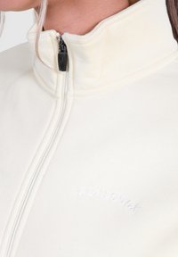 Krämfärgad zip-up jacka med hög krage, mjuk textur och broderad logotyp framtill. Har en kontrasterande svart dragkedja.