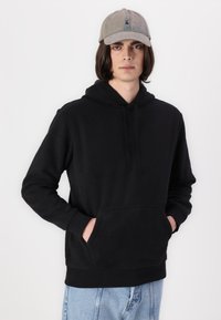 Hanorac negru din fleece cu buzunar frontal și șnururi ajustabile, asociat cu o șapcă gri cu un logo simplu. Blugi albaștri completează ținuta.