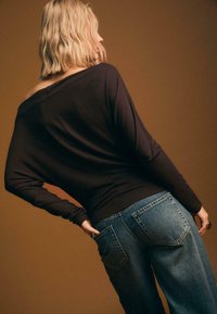 Top lungo con maniche, senza spalle, di colore marrone scuro abbinato a jeans a gamba larga blu chiaro. Tessuto morbido, vestibilità rilassata e dettagli classici con tasche.
