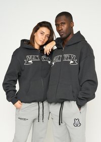 Dos personas llevan ponchos a juego de color gris oscuro con cremallera que presentan la palabra "MULTIPLY" bordada en blanco, combinados con pantalones de chándal gris claro.