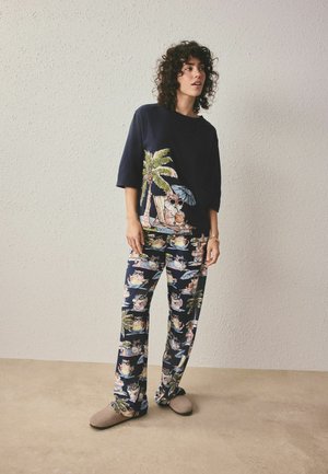 Jonge vrouw in marineblauwe pyjama met print van kat en palmboom, staand tegen een licht gestructureerde muur en beige slippers dragend.